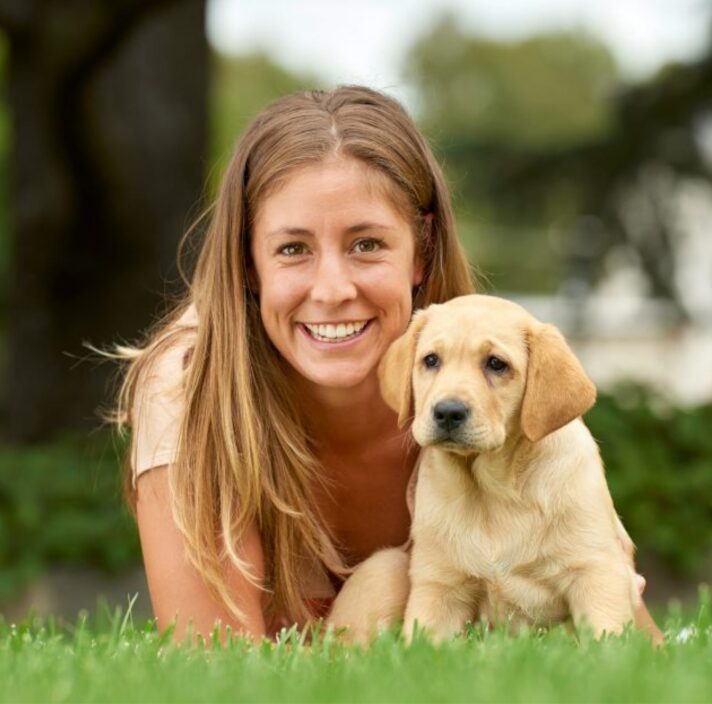 A person laying on some grass with an eight week old yellow labrador puppy. The person is smiling and looking at the camera. The puppy is also looking at the camera.
