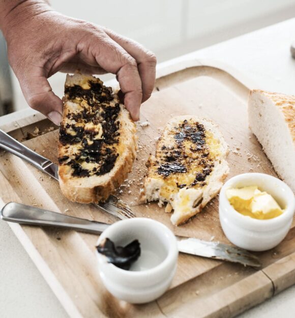 A person picking up a piece of buttered toast that is on a wooden board. There is a second piece of toast on the board